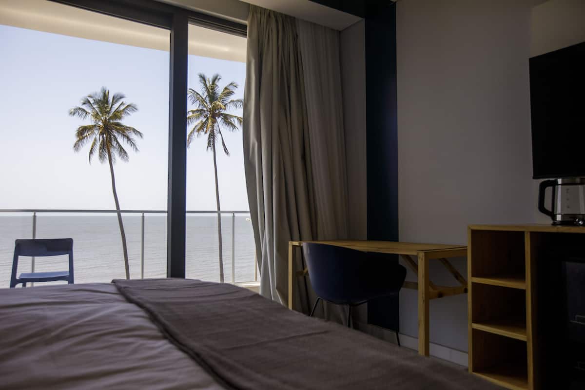 A hotel room with a bed, a desk, a chair, a TV, and a view of the ocean with palm trees.