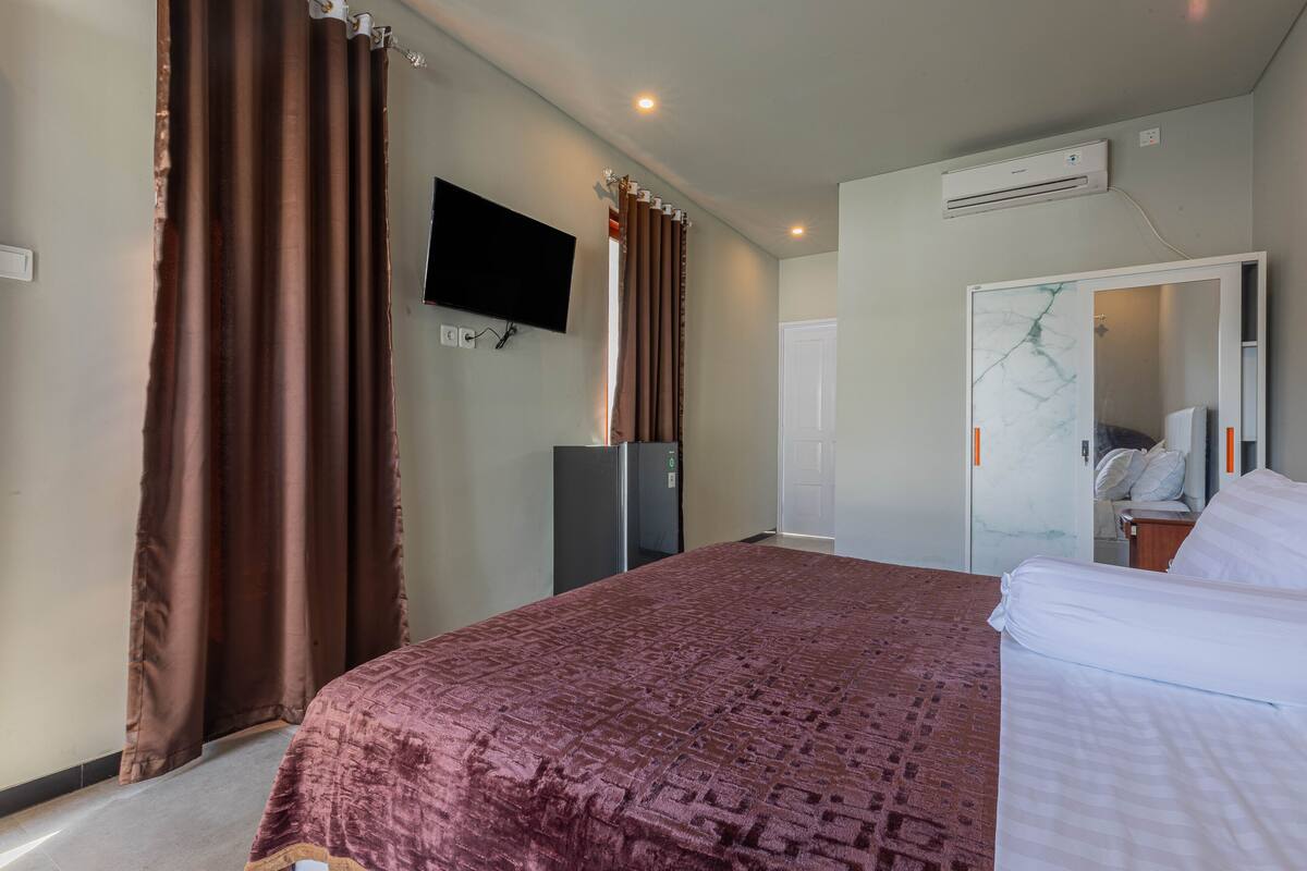 A hotel room with a large bed, a flat-screen TV mounted on the wall, a wardrobe, and an air conditioning unit.