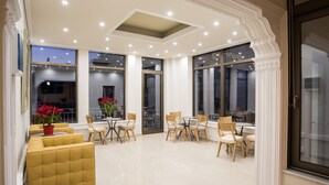 Una habitación amplia con techo alto, ventanas grandes y una zona de estar central con un sofá amarillo y sillas de madera.