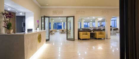 A modern hotel lobby with a reception desk, seating area, and glass doors leading to a dining area.