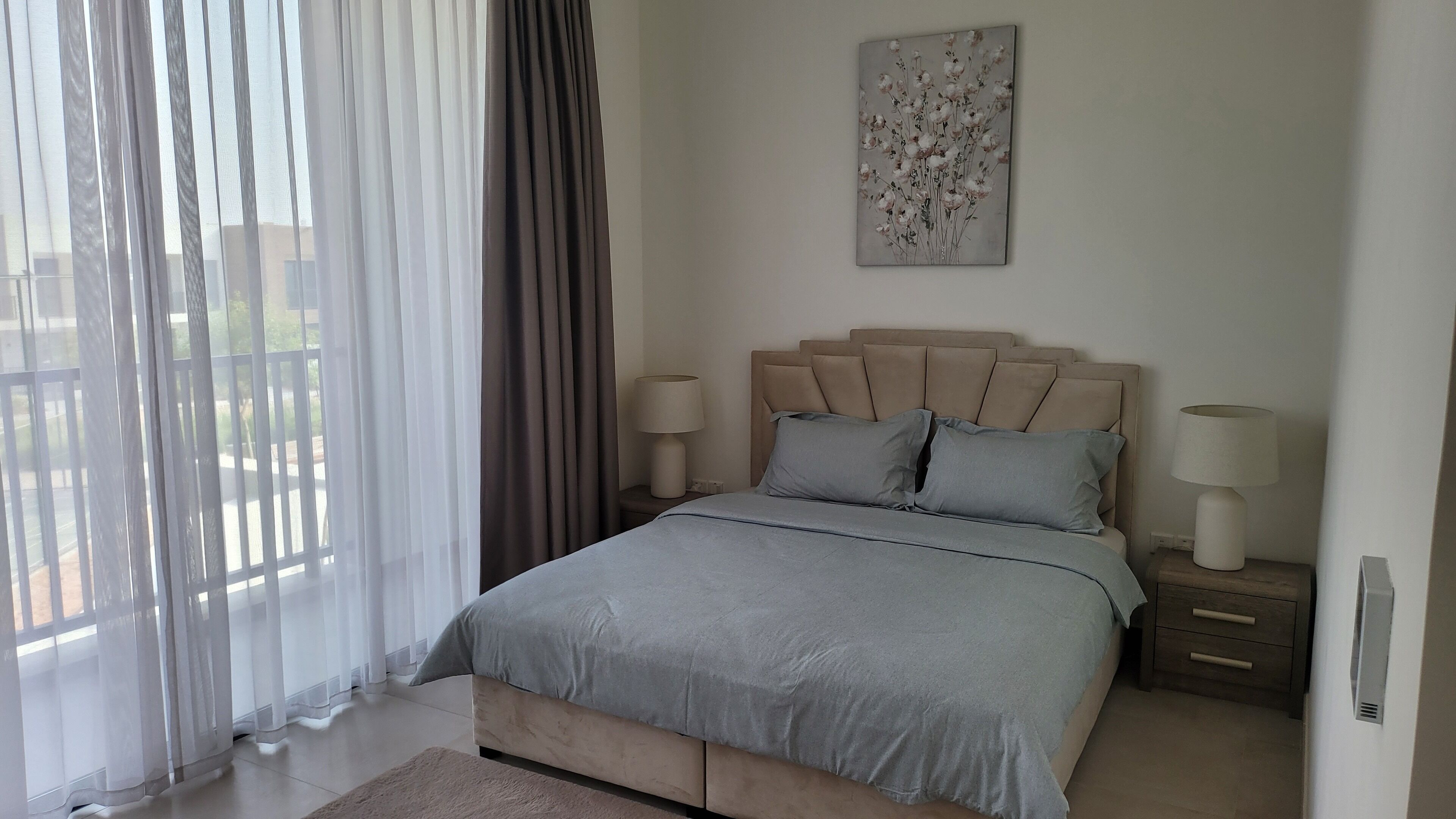 A bedroom with a bed, two bedside tables, a nightstand, a window with sheer curtains, and a wall-mounted artwork.