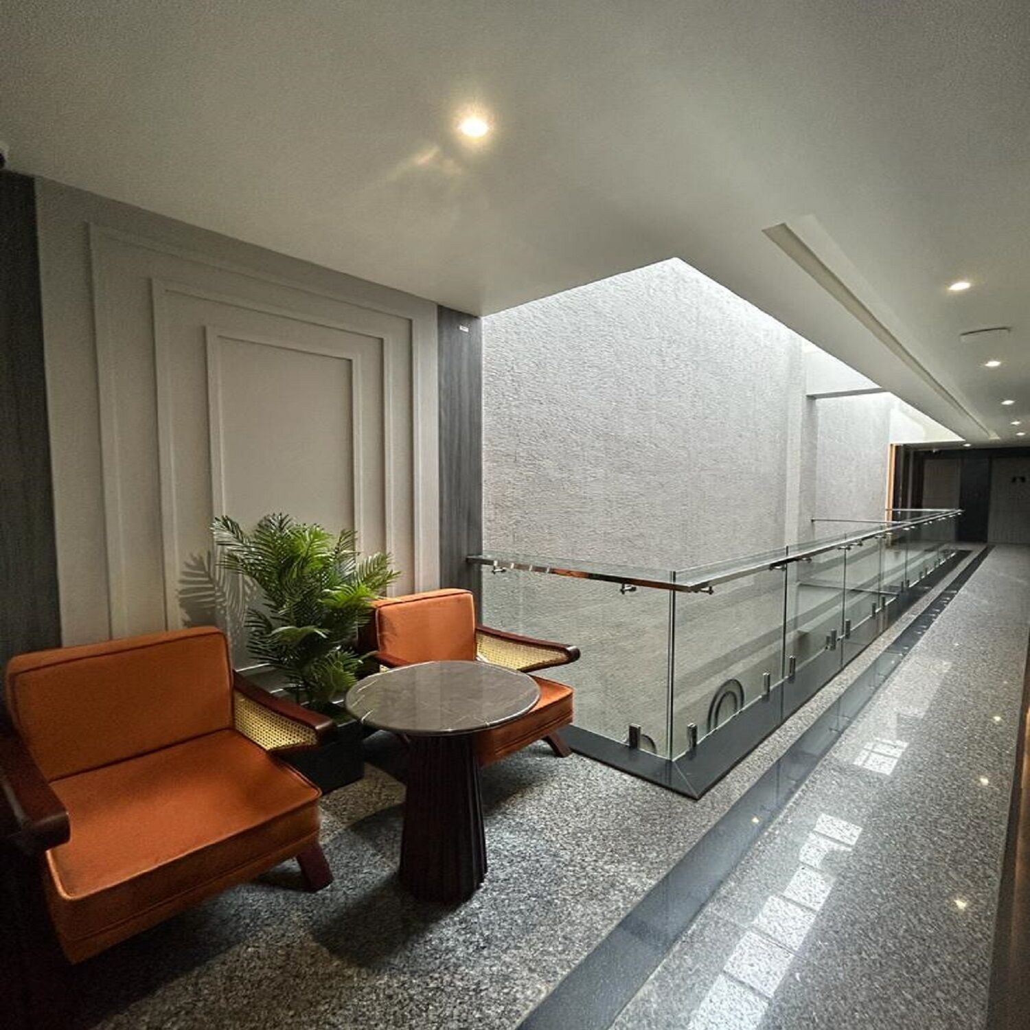 A modern lobby with a seating area featuring orange chairs and a round table, a glass railing, and a large window providing natural light.