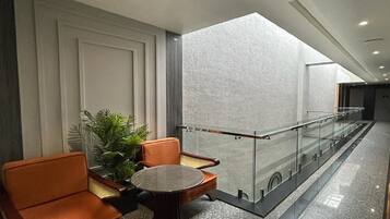 Een moderne lobby met een zithoek, met oranje stoelen en een ronde tafel, een glazen balustrade, en een groot raam dat voor natuurlijk licht zorgt.