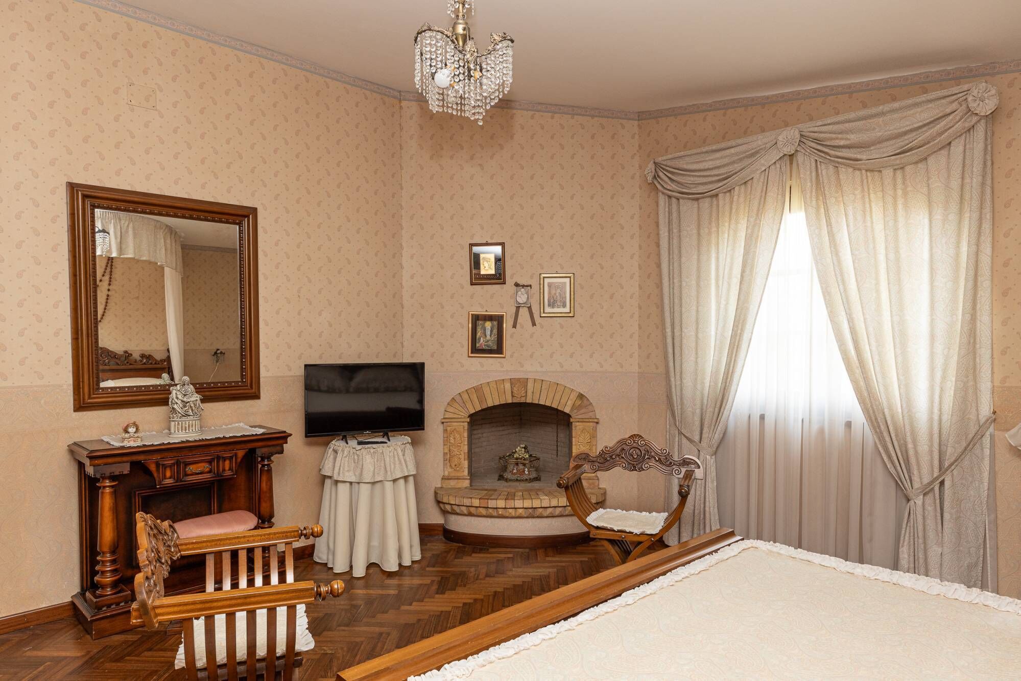 A room with a wooden floor, a fireplace, a television, a bed, a chair, a table, a mirror, and curtains.