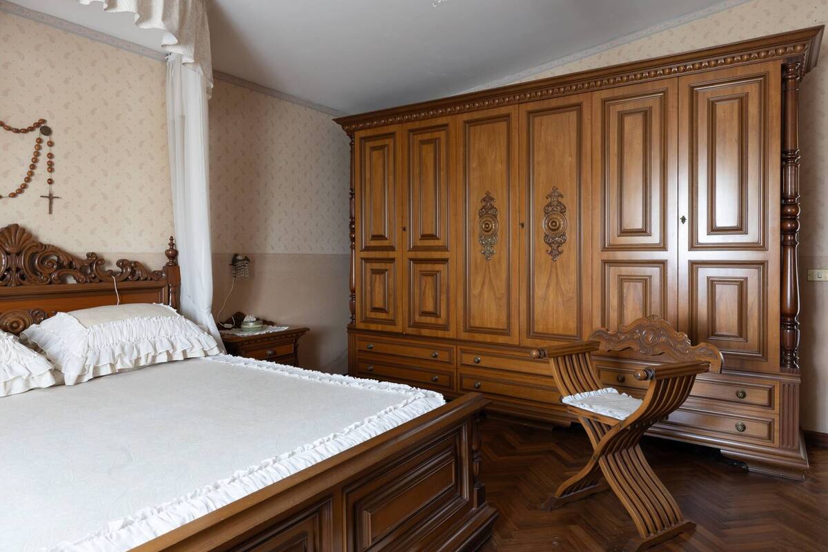 Een slaapkamer met een grote houten garderobe, een houten stoel met een bekleed zitvlak en een bed met wit beddengoed.