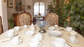 Een eettafel gedekt met witte kopjes, schoteltjes en een kristallen kom, omringd door stoelen met bloemenmotieven.