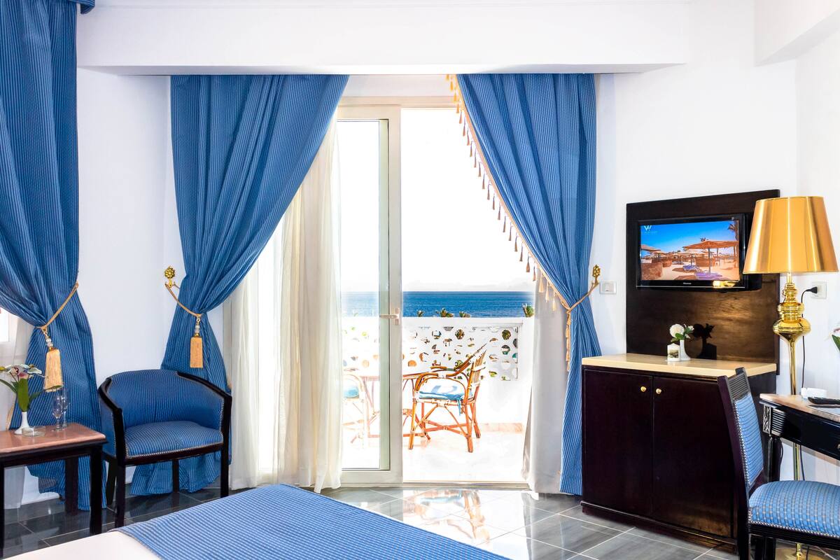 A hotel room with a bed, a desk, a TV, and a view of the sea through the glass door.