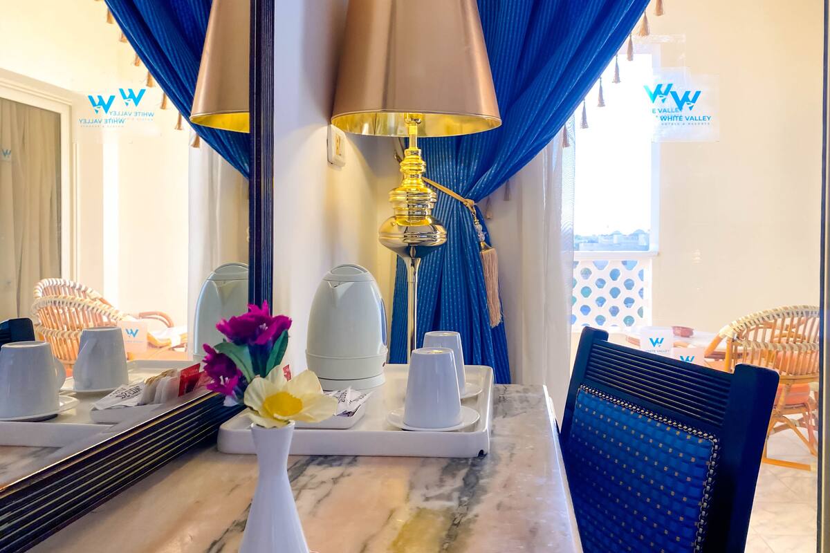 A hotel room with a marble table, a white vase with flowers, a tray with white cups, a lamp, and blue curtains.