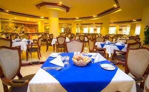 Un comedor con varias mesas dispuestas para recibir a los comensales, con mantel azul, individuales blancos y diferentes tipos de vajilla, incluyendo platos, vasos y cestas de pan.