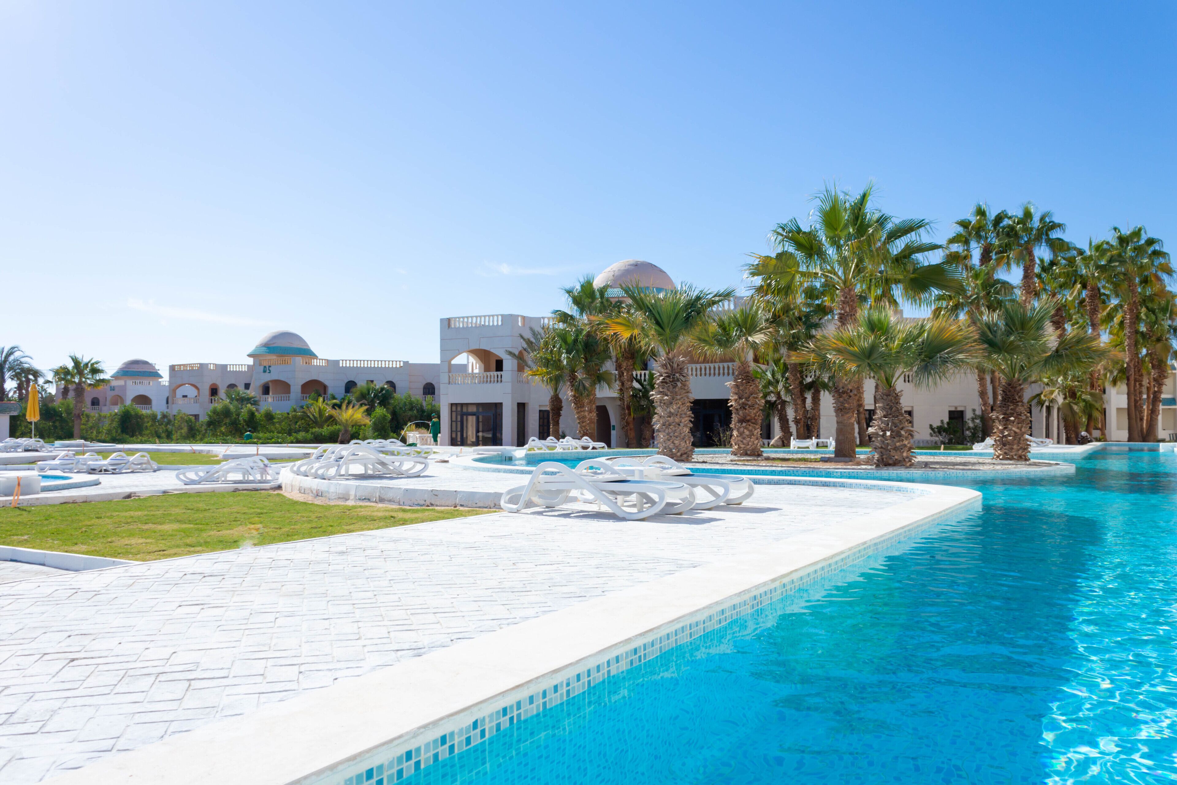 Zona de piscina con palmeras, sillas de descanso y un edificio con techos abovedados al fondo.