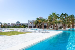 Zona de piscina con palmeras, sillas de descanso y un edificio con techos abovedados al fondo.