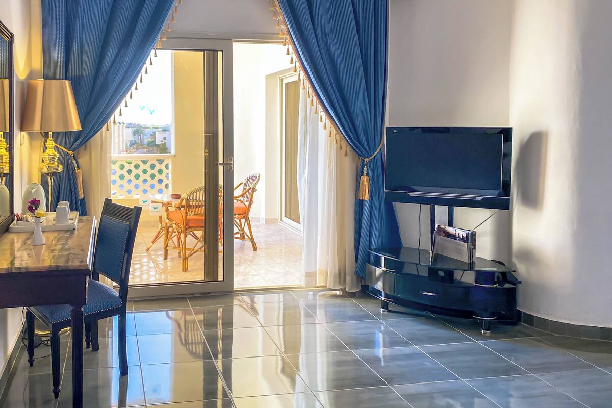 A modern living room with a glass door leading to a balcony, a dining area with wicker chairs, and a flat-screen TV mounted on the wall.