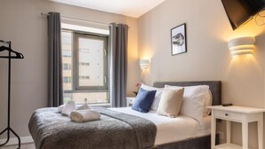 A modern bedroom with a large bed, a bedside table, a TV mounted on the wall, and a window with curtains. - Belfast Quarters: Modern City Centre Hub 3 Bed! (Belfast)