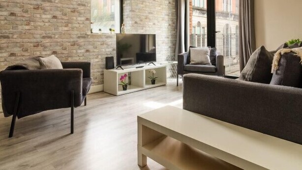 A modern living room with a brick wall, a flat-screen TV, two sofas, and a wooden coffee table. - Belfast Quarters: Modern City Centre Hub 3 Bed! (Belfast)