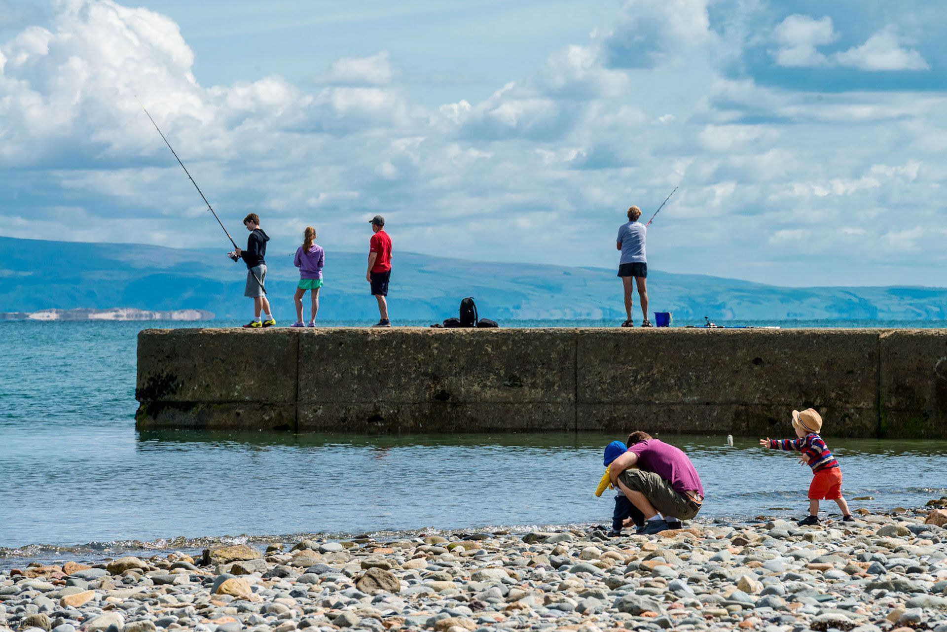 A group of people fishing by a rocky shore with a view of the sea and distant mountains.