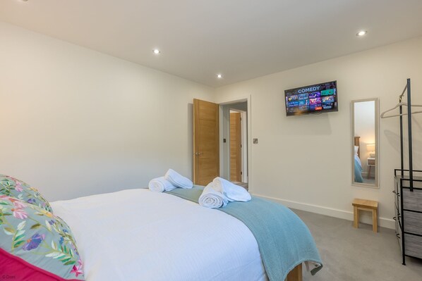 Una habitación de hotel con una cama, un televisor montado en la pared mostrando un canal de comedia, una puerta de madera, un taburete pequeño de madera y un armario metálico.