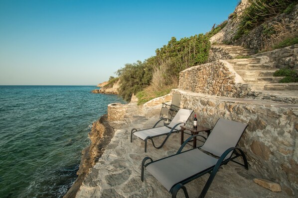 A seaside terrace with stone steps leading down to the water, equipped with lounge chairs and a table.