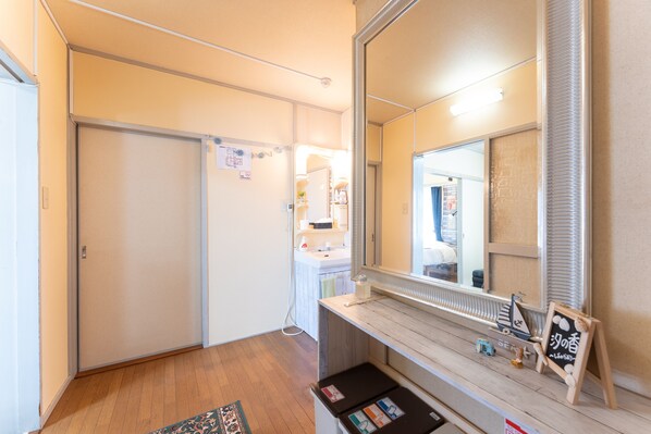Quarto de hotel com piso de madeira, um espelho grande, uma pequena mesa com uma placa e uma porta que dá para outro quarto.