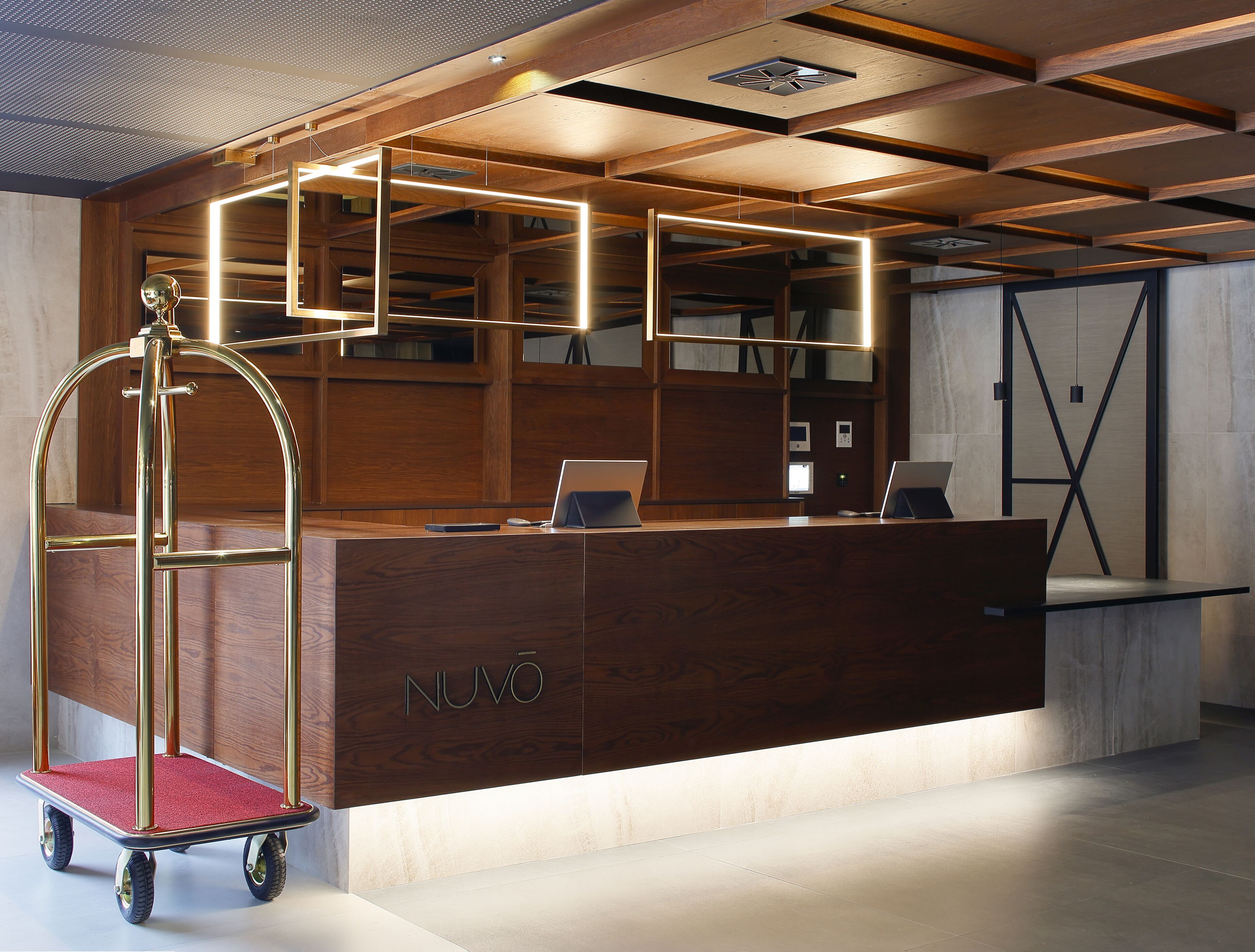 Eine Rezeptionstheke mit Holzoptik und dem Schriftzug "NUVO". Ein rollbarer Wagen mit goldfarbigem Griff und einer roten Matte. Die Decke zeigt ein Holzdesign mit sichtbaren Balken und einer zentralen Leuchte.
