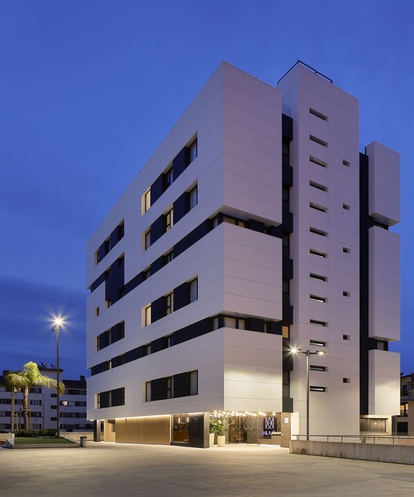 A multi-story building with a modern architectural design, featuring a flat facade, multiple windows, and a ground-level entrance with a well-lit entrance area. - NUVÓ HOTEL BOUTIQUE (Oviedo)