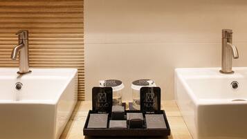 Two sinks with modern faucets, a wooden panel wall, and a tray with body milk bottles and boxes.