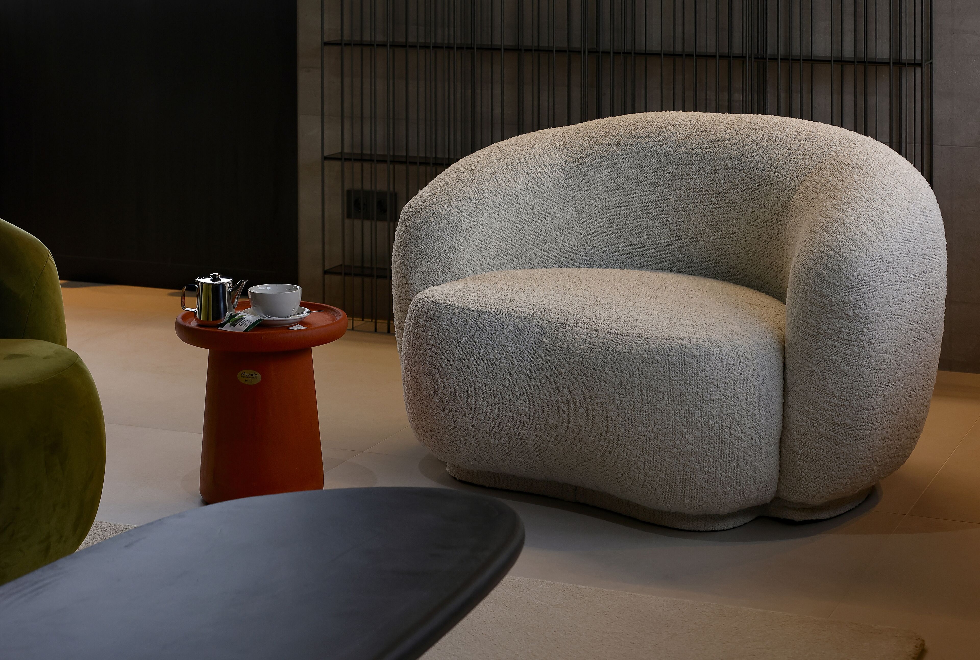 Una habitación moderna con un sillón de diseño redondeado, una pequeña mesa roja con una bandeja que sostiene una taza y una tetera, y un sillón verde.