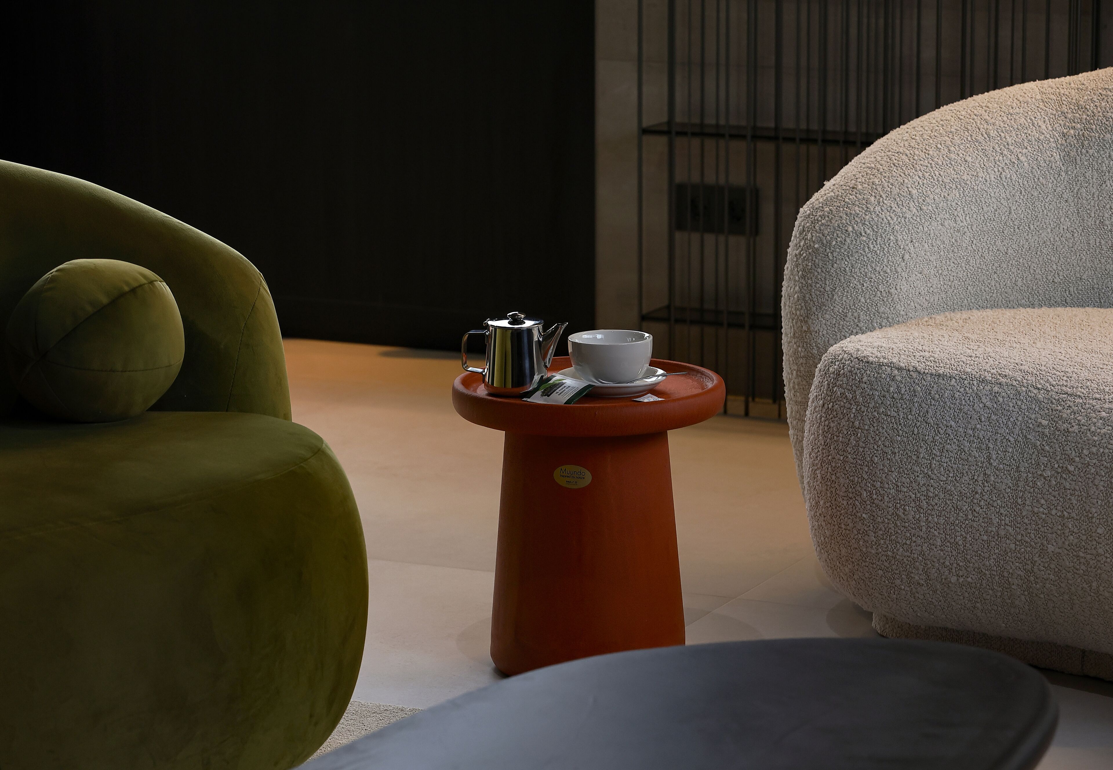 A modern room with a green sofa, a white armchair, and a small orange table with a white cup and a silver teapot.