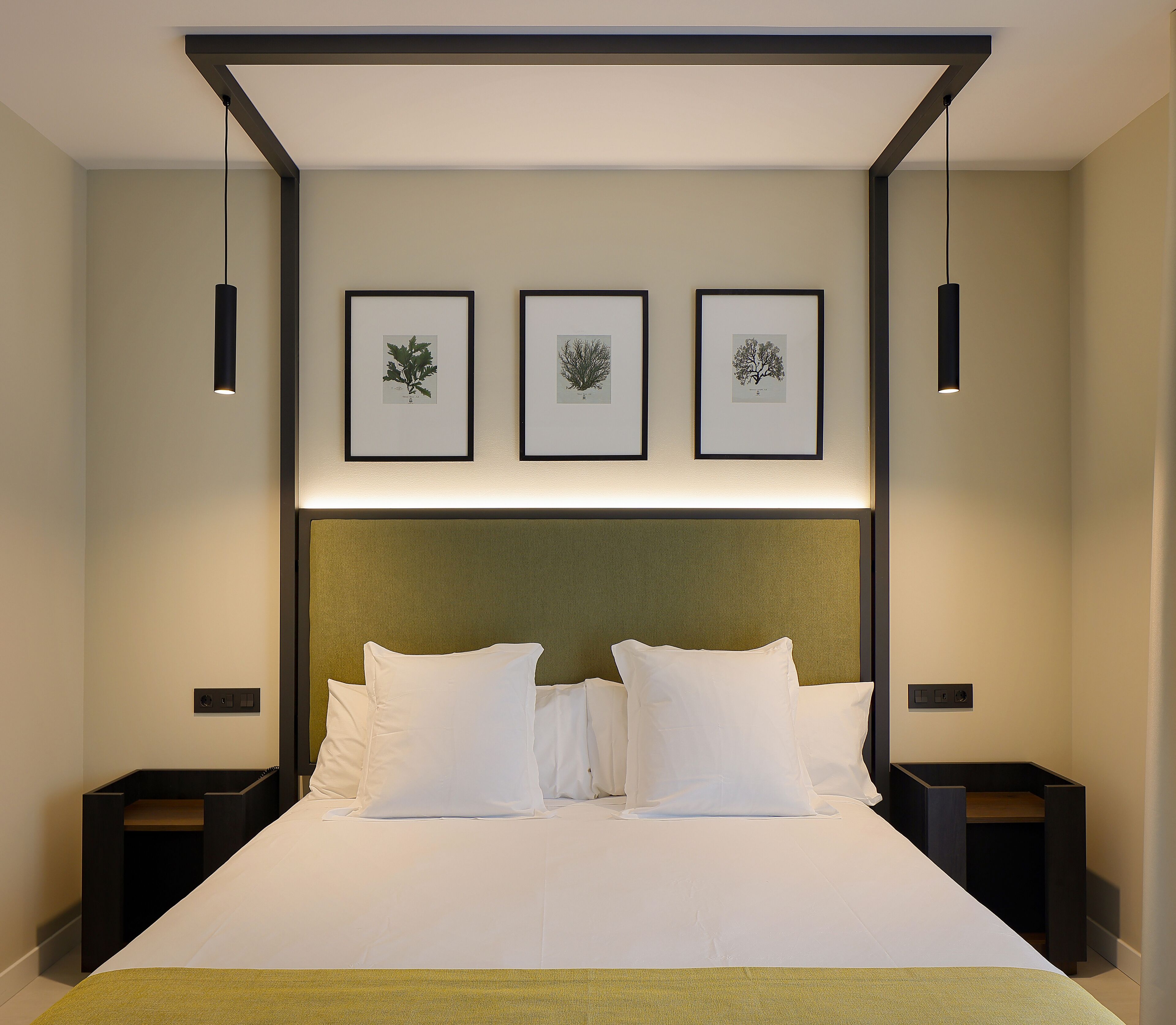 Habitación de hotel con una cama grande, tres láminas botánicas enmarcadas en la pared y dos mesitas de noche con lámparas.