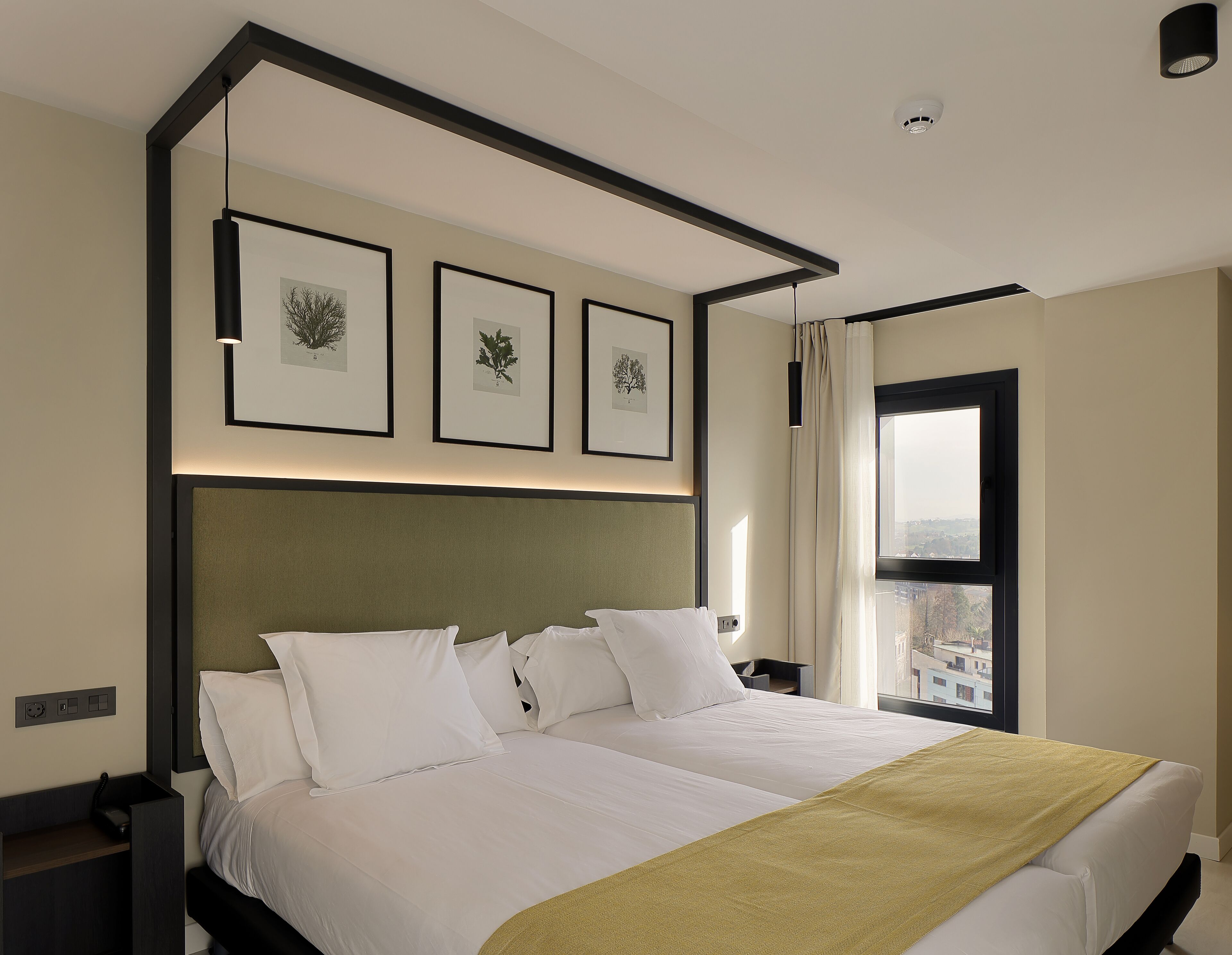 A hotel room with a large bed, a headboard, a window with curtains, and framed botanical prints on the wall.
