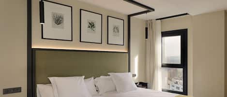A hotel room with a large bed, a headboard, a window with curtains, and framed botanical prints on the wall.