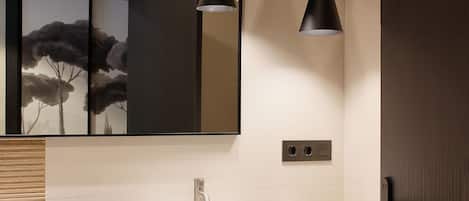 A modern bathroom with a large mirror, a white sink, black pendant lights, and a small potted plant.