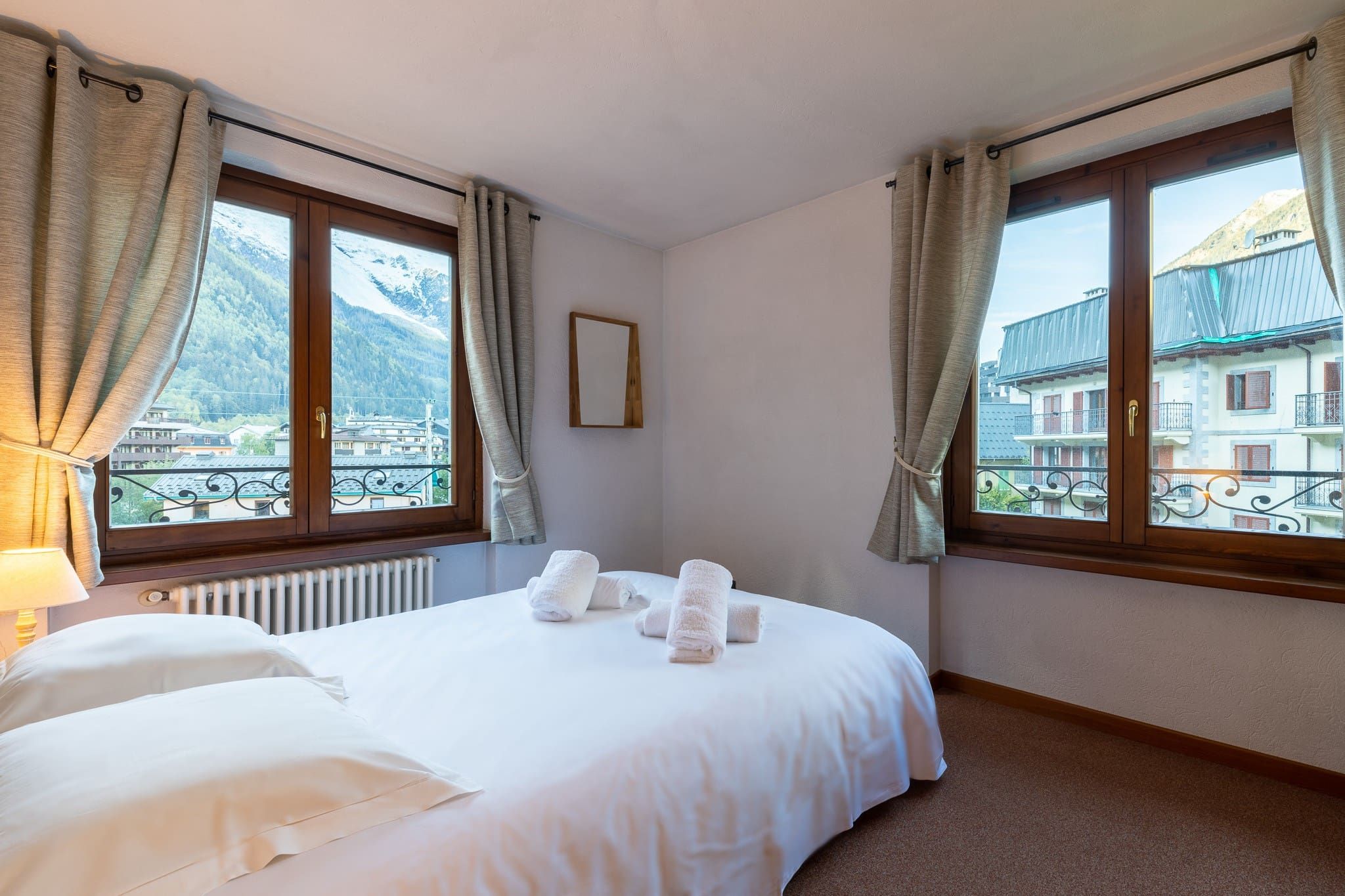 A hotel room with a bed, two windows with curtains, a radiator, and a view of buildings and mountains.