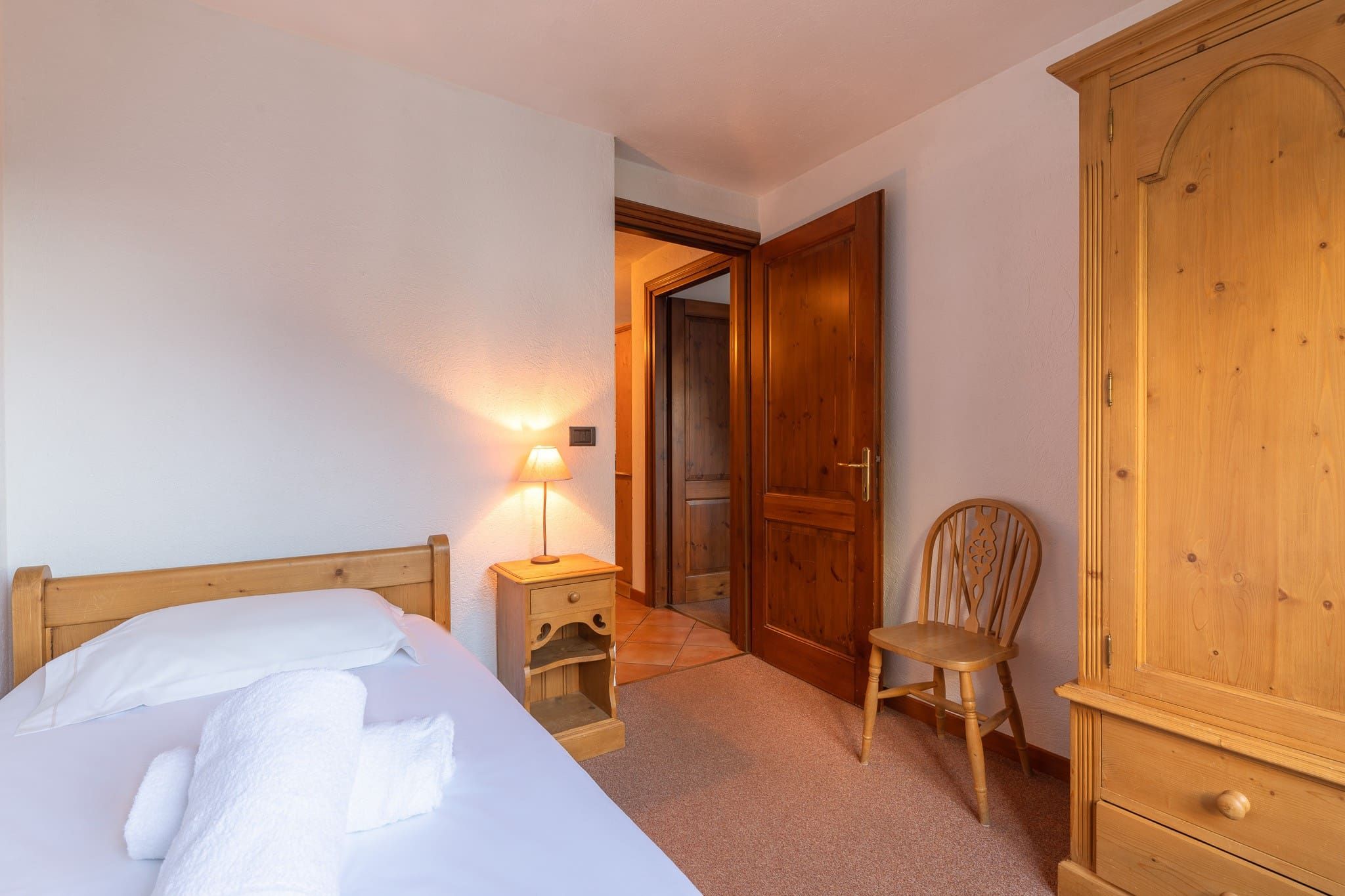 A hotel room with a bed, a nightstand with a lamp, a wooden wardrobe, and a chair.