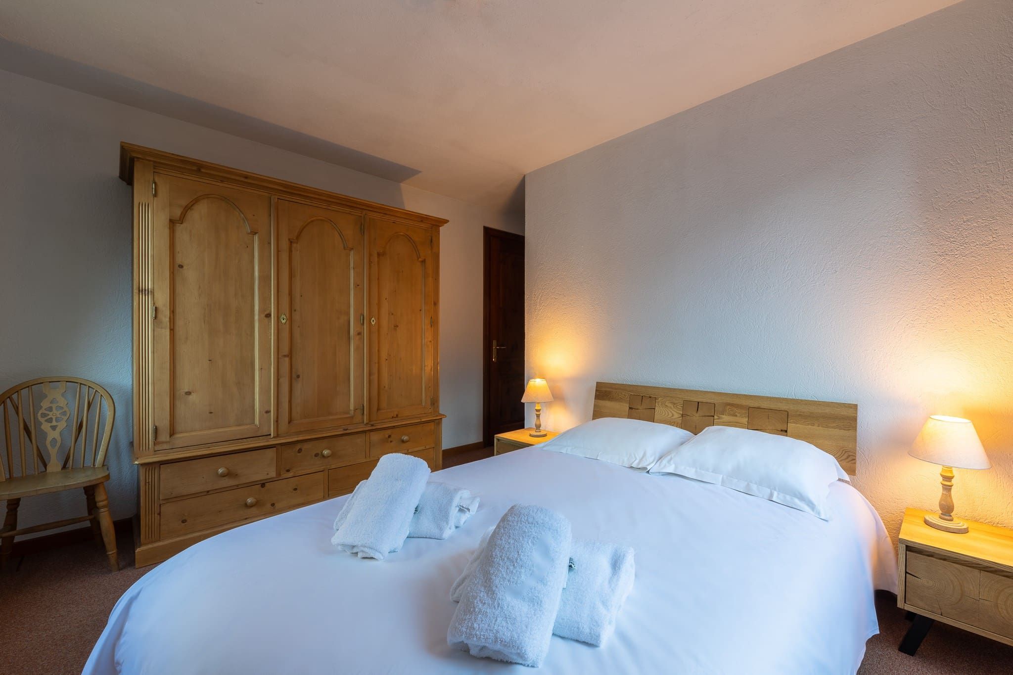 A hotel room with a large bed, two bedside lamps, a wooden wardrobe, and a chair.