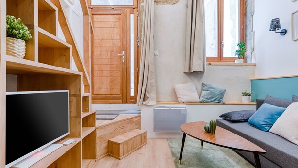 A modern living room with a flat-screen TV, wooden shelving, a sofa with cushions, a small table, and a window with curtains. - Smartstay Louis Chazette - Centre-ville (Lyon)