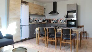 A modern kitchen with wooden cabinets, a central island with chairs, a dining table, and a built-in oven. - Gîte Padura - Location 3 Chambres - Braud et Saint Louis (Braud-et-Saint-Louis)