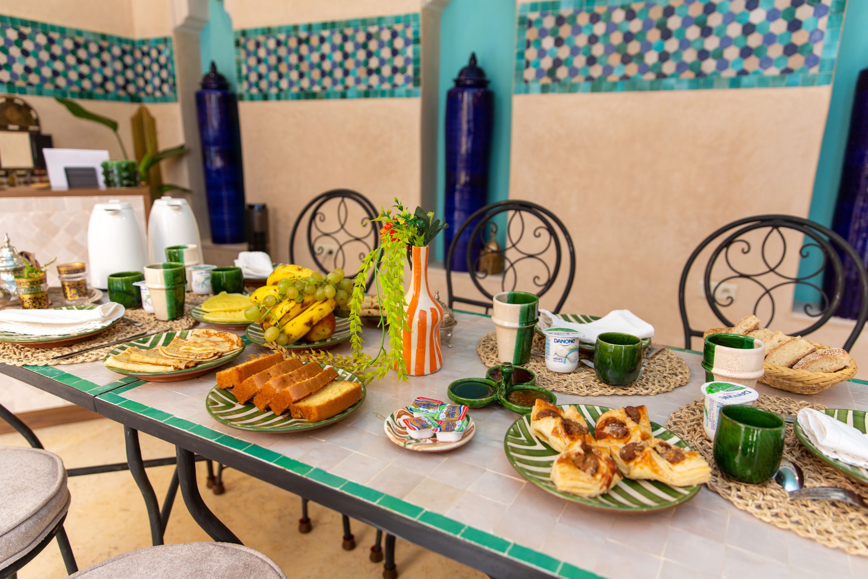 Una mesa servida con diversos platos, incluyendo frutas, pan y pasteles, en un comedor con baldosas estampadas y sillas de metal.