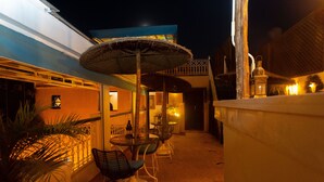 Un patio al aire libre con una mesa redonda y sillas, un paraguas de paja y un pequeño jardín con una planta de palmera.
