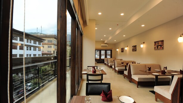 A restaurant with a view of buildings through large windows. Tables are set with plates, glasses, and napkins. There are cushioned seating areas and wall-mounted lights. - Thimphu Central Hotel (Thimphu)
