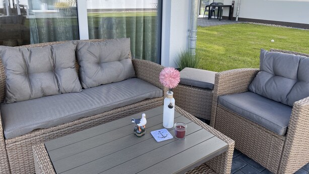Un conjunto de sofá y sillón de mimbre con cojines, una mesita con una vela y una figurita de pájaro, y vistas a un jardín césped y un edificio al fondo.