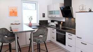 Una cocina moderna con armarios blancos, un horno negro y una encimera de acero inoxidable. Hay un taburete de bar y un florero con una planta sobre la encimera.