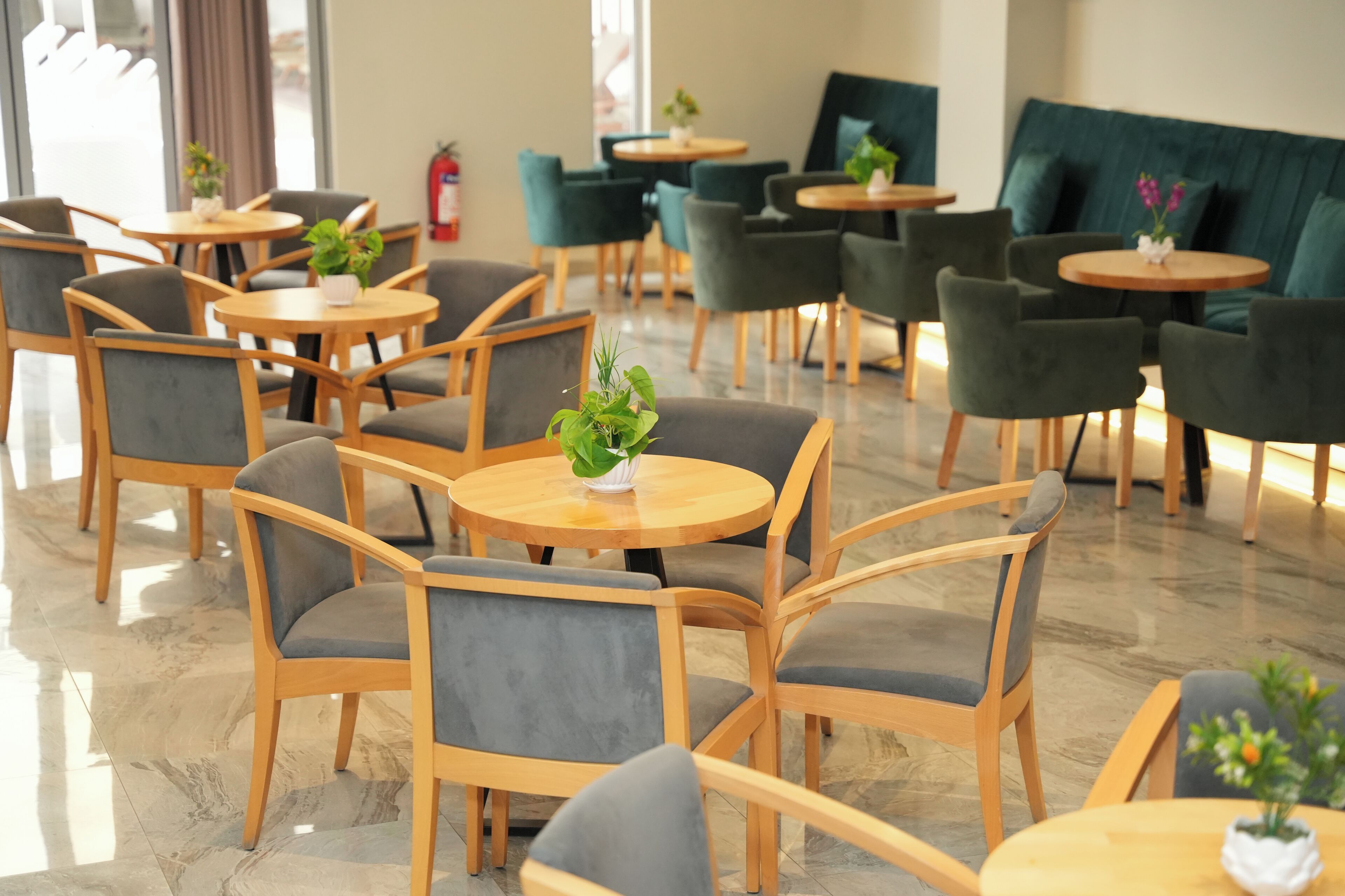 A hotel lobby with round tables and chairs, some with plants. There are green sofas and a fire extinguisher on the wall.