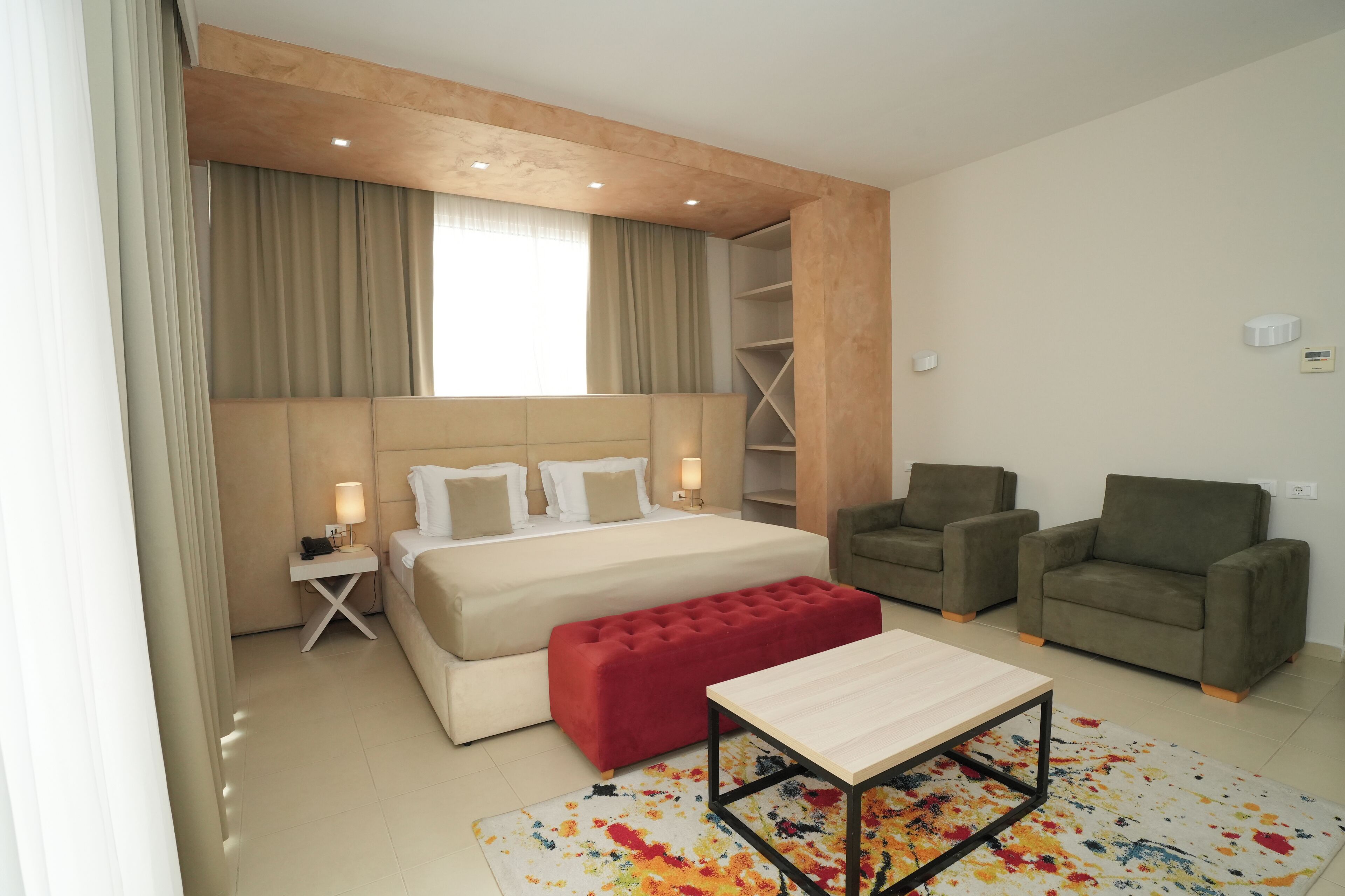 A modern hotel room with a large bed, two armchairs, a red bench, a small table, and a colorful rug.