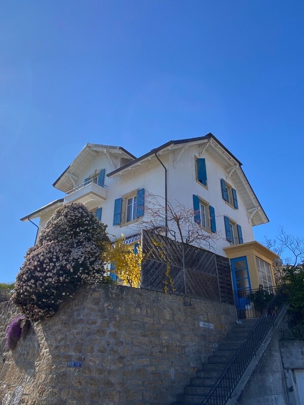 Un bâtiment à deux étages, avec un mur de pierre à l’avant, un escalier sur le côté droit et un ciel bleu dégagé.