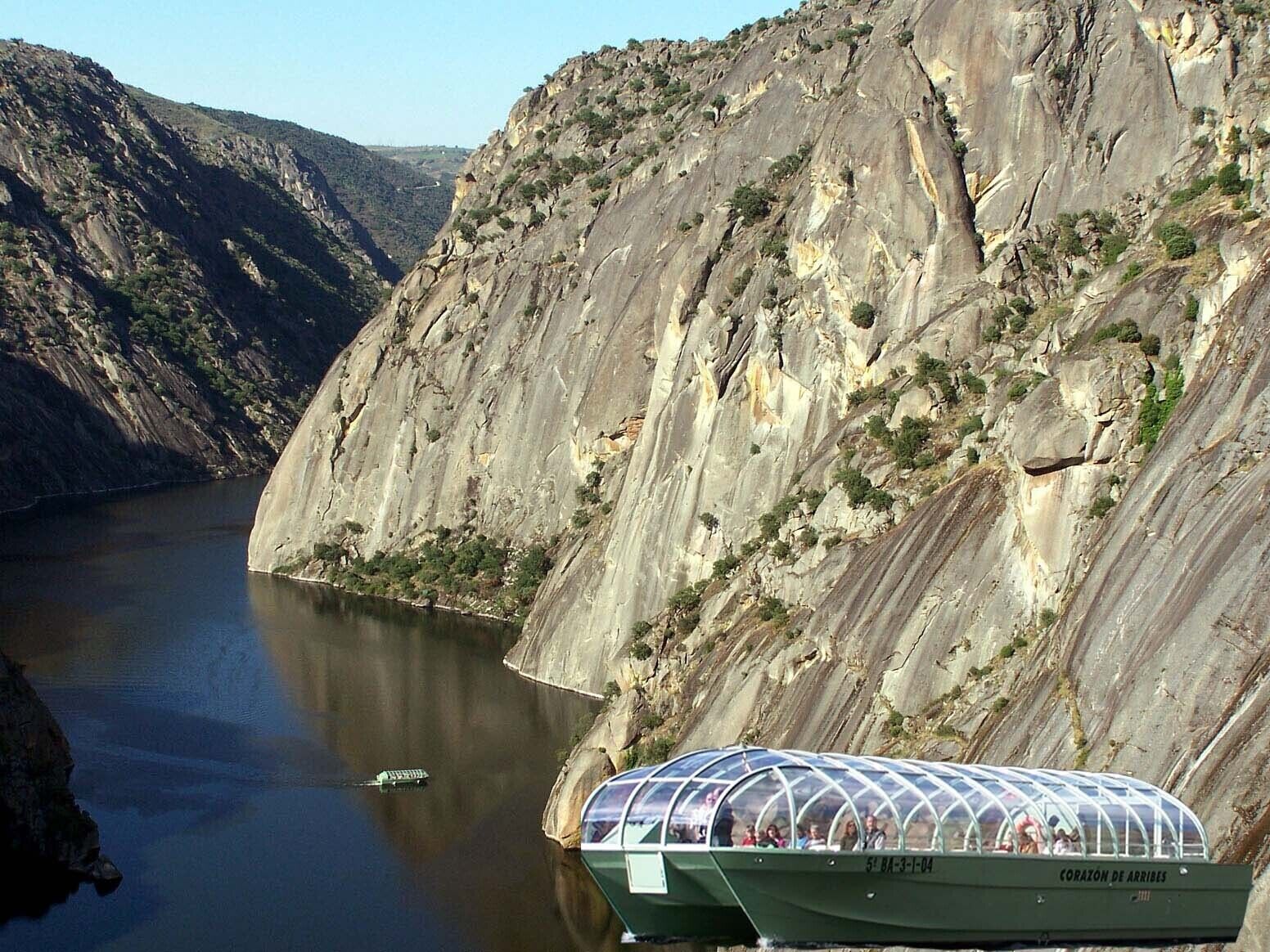 Een boot met een transparant dak, voorzien van de tekst 'CORAZON DE ARRIVES', afgemeerd aan een rivier met hoge, rotsachtige kliffen op de achtergrond.