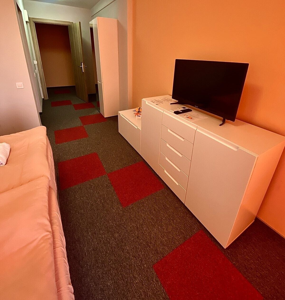 Een hotelkamer met een bed, een televisie op een dressoir en een vloerkleed met rode en grijze vierkanten.