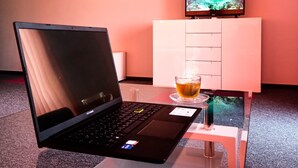 A laptop on a glass table with a cup of tea, a white cabinet, and a TV in the background. - Ubis hotel (Rodopi)