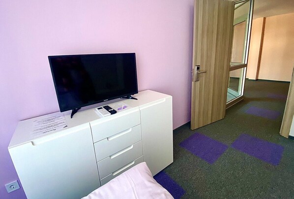 A room with a white desk, a flat-screen TV, a small table with a remote and a pen, and a door leading to another room. - Ubis hotel (Rodopi)