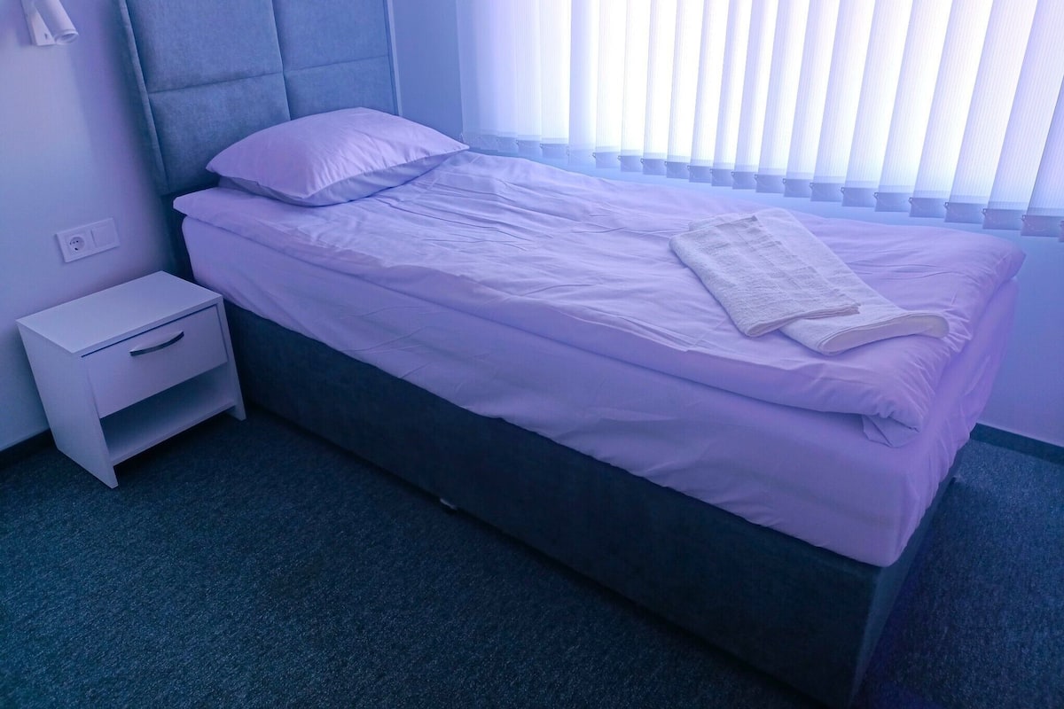 A neatly made bed with a pillow and a folded towel, a small bedside table with a drawer, and a power outlet on the wall.