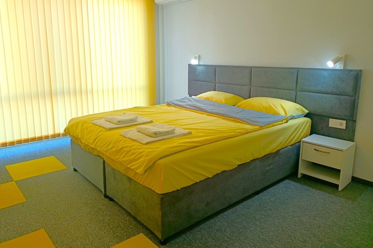 A hotel room with a bed, a nightstand, and a window with blinds.